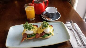 En frukosttallrik med ägg Benedict, en röd vattenkokare, ett glas apelsinjuice och en kopp på ett träbord.