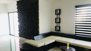 A modern living room with a sectional sofa, a round black ottoman, a small side table, and a wall-mounted shelf. - Apartamento 1 (Cancun)