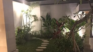 Un sendero en un jardín con piedras que guían a través de abundante vegetación, rodeado de diversas plantas y una edificación.