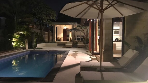 Zona de piscina nocturna con piscina, sillas de descanso y una sombrilla.