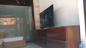 Una habitación con una televisión sobre un soporte de madera, un sofá de mimbre y un ventanal de vidrio que da al exterior.