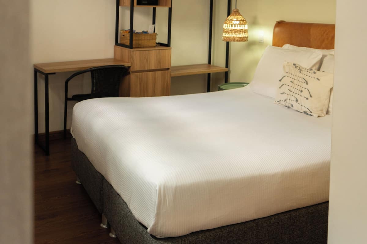A bedroom with a bed, a desk, a chair, a hanging light fixture, and a wall-mounted shelf.