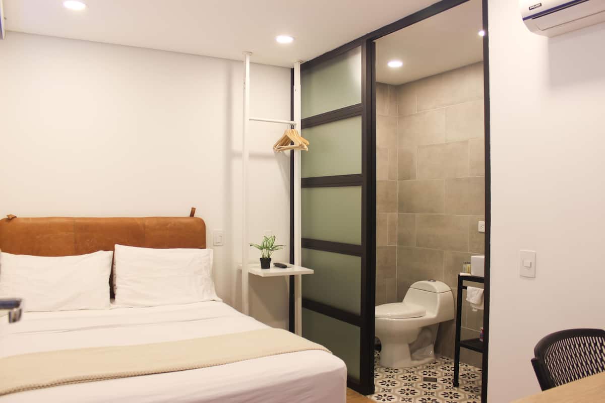 A hotel room with a bed, a small bedside table with a plant, a toilet, and a chair.