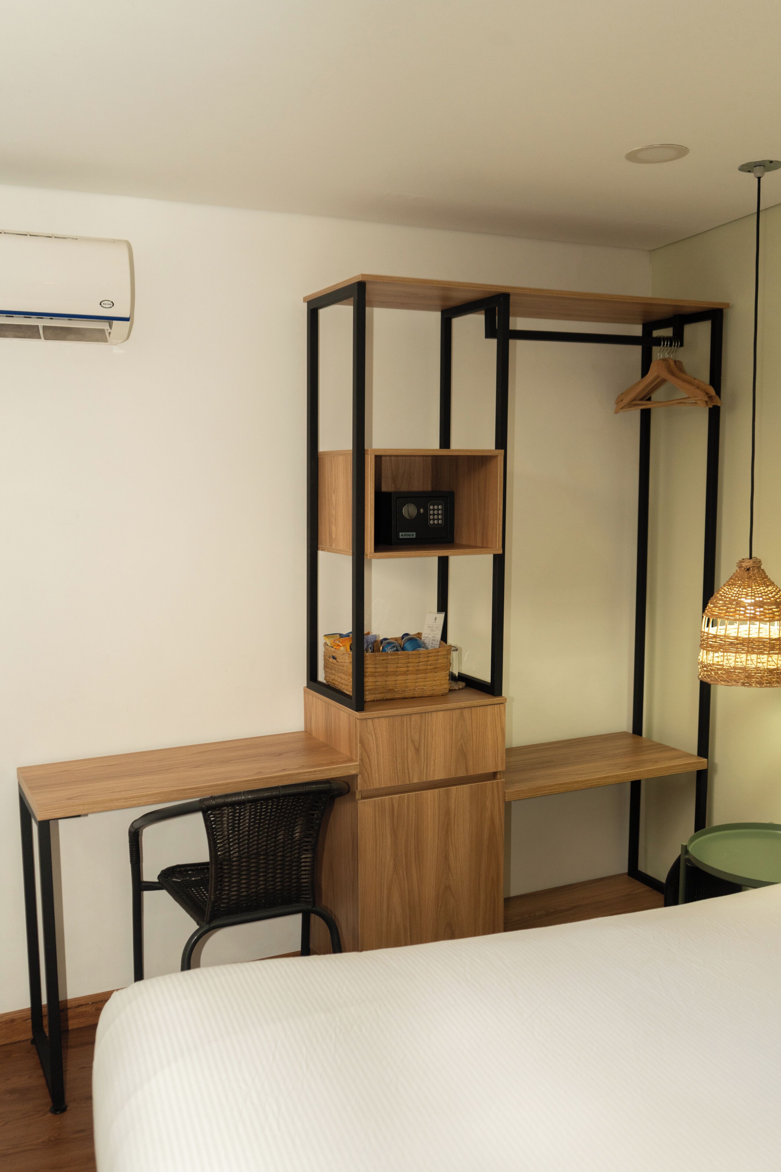 A bedroom with a wooden desk, a chair, a bed, a hanging light fixture, and a wall-mounted air conditioner.