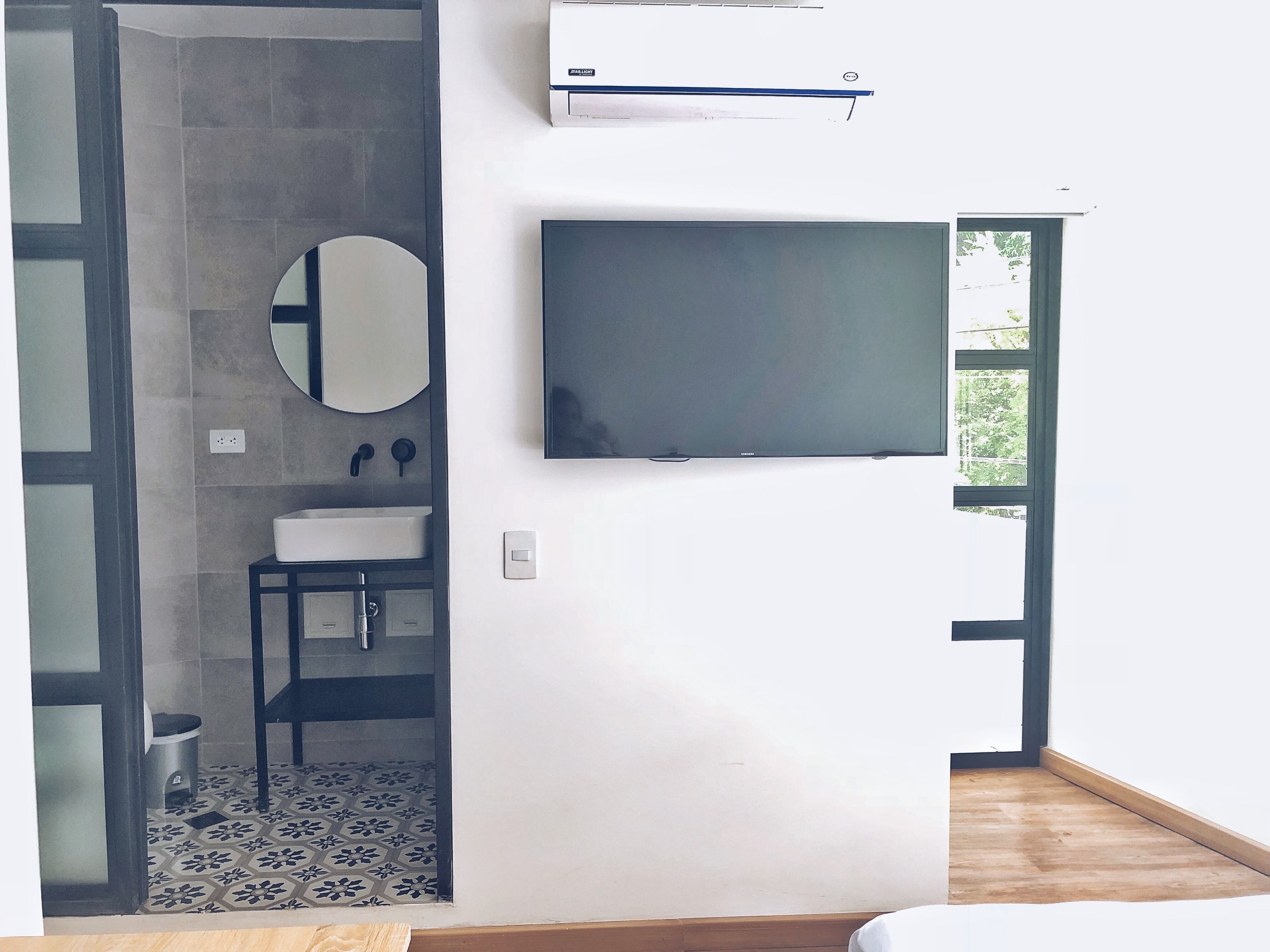 Un baño moderno con ducha, lavamanos e inodoro. Hay un televisor de pantalla plana montado en la pared y una unidad de aire acondicionado encima.