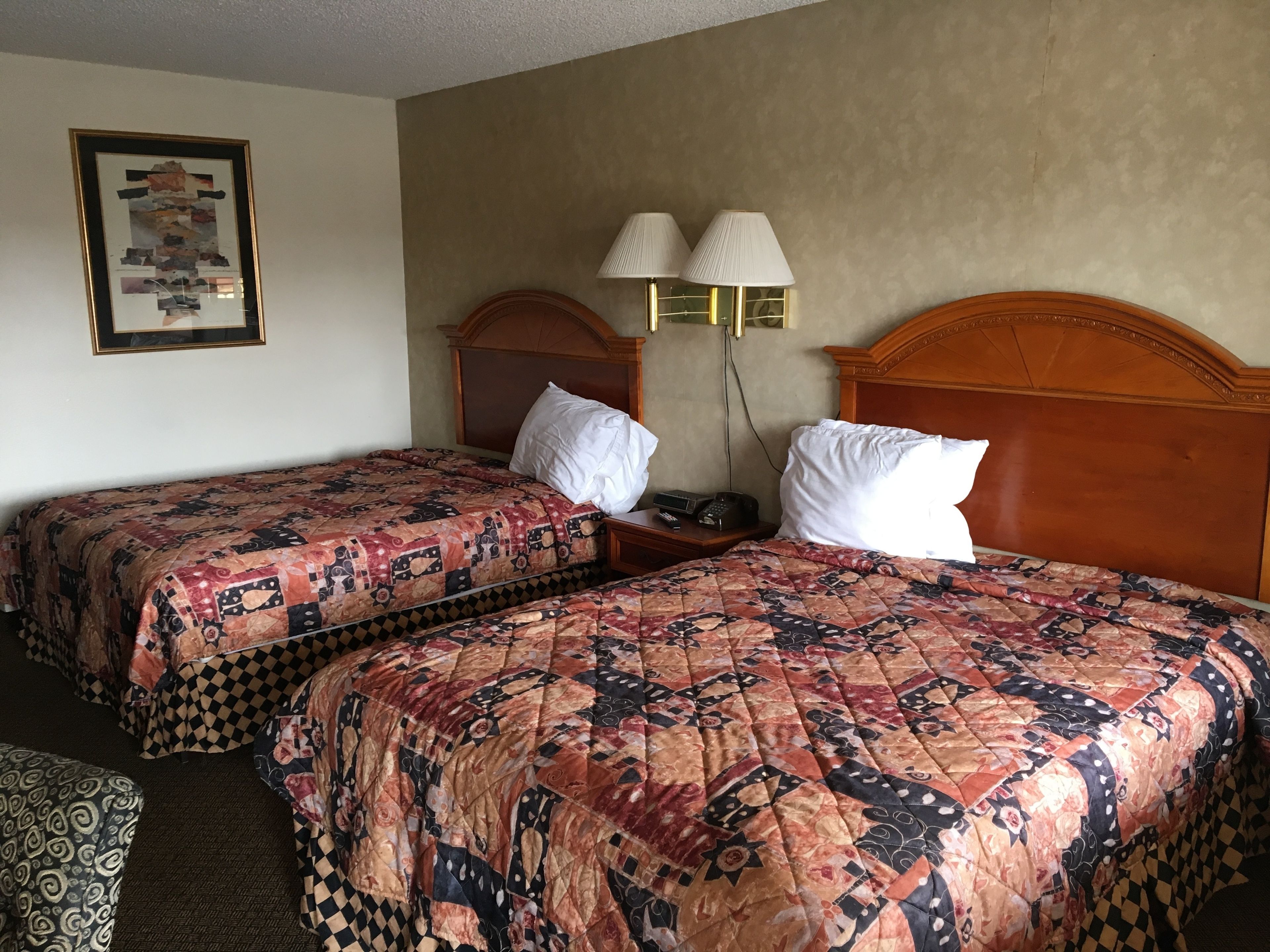 A hotel room with two beds, each with a patterned quilt, a wooden headboard, bedside lamps, and a framed picture on the wall.