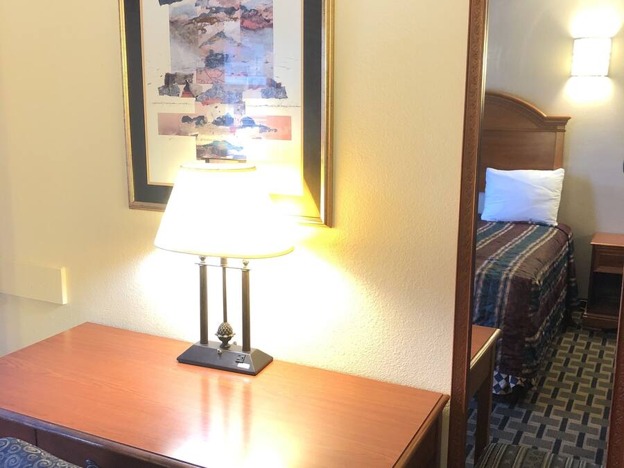 A hotel room with a wooden desk, a lamp, a framed abstract artwork, a large mirror, a bed with a patterned blanket, and a patterned rug.