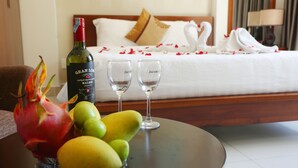Une chambre d’hôtel avec un lit, une bouteille de vin, deux verres et un panier de fruits sur une table.