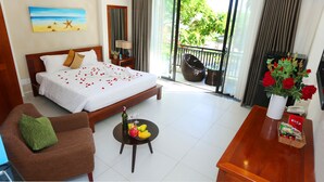 Une chambre d’hôtel avec un lit, une chaise, une table avec des fruits, un vase de fleurs et un balcon donnant sur un paysage verdoyant.