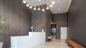 Zona de recepción con un mostrador blanco, una recepcionista, una planta en maceta, un banco y una puerta al fondo.