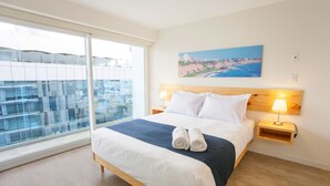 Habitación de hotel con una cama grande, un cuadro de una escena costera, una ventana con vista a edificios y una mesita de noche de madera con una lámpara.