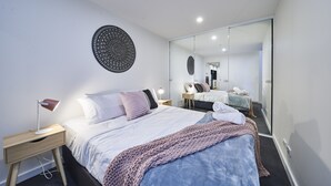 Quarto moderno com uma cama grande, uma mesa de cabeceira com luminária, uma obra de arte circular na parede e um espelho refletindo o ambiente.