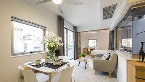 Una habitación de hotel moderna con zona de comedor, sofá, una mesita con una planta y ventana con vista a la ciudad.