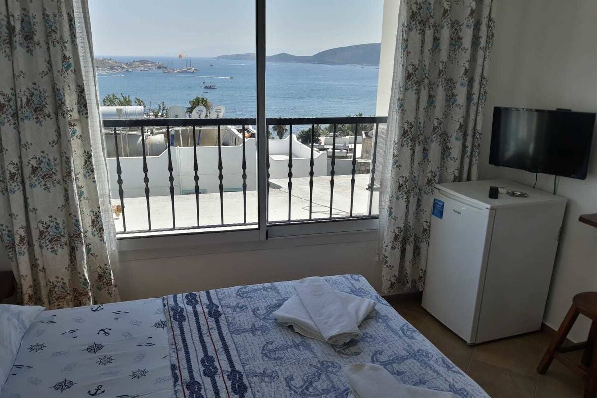 Quarto de hotel com cama, pequeno frigorífico, televisão e varanda com vista para o mar e barcos.