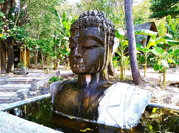 Une statue de Bouddha se trouve dans un bassin, entourée d’une végétation luxuriante et de structures en pierre.