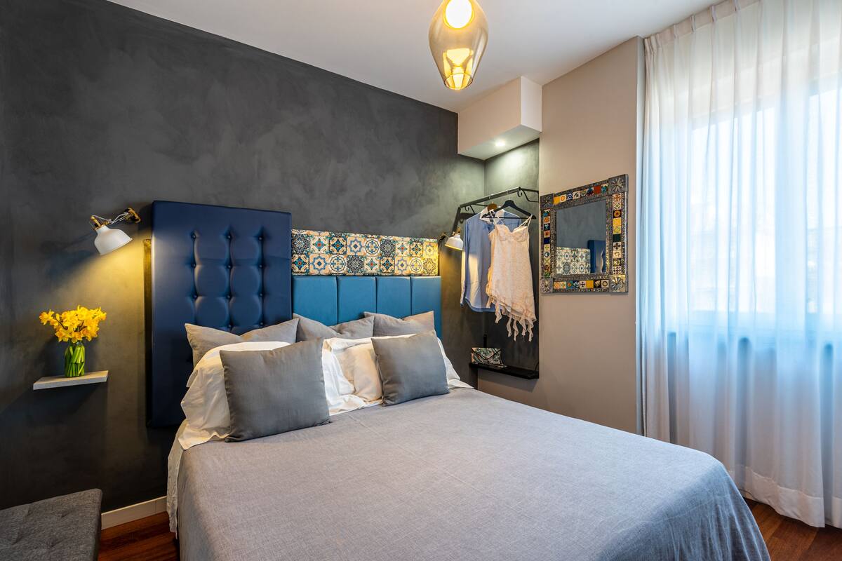 A modern bedroom with a tufted headboard, a bed with grey linens, a bedside table with a vase of flowers, and a hanging garment rack.
