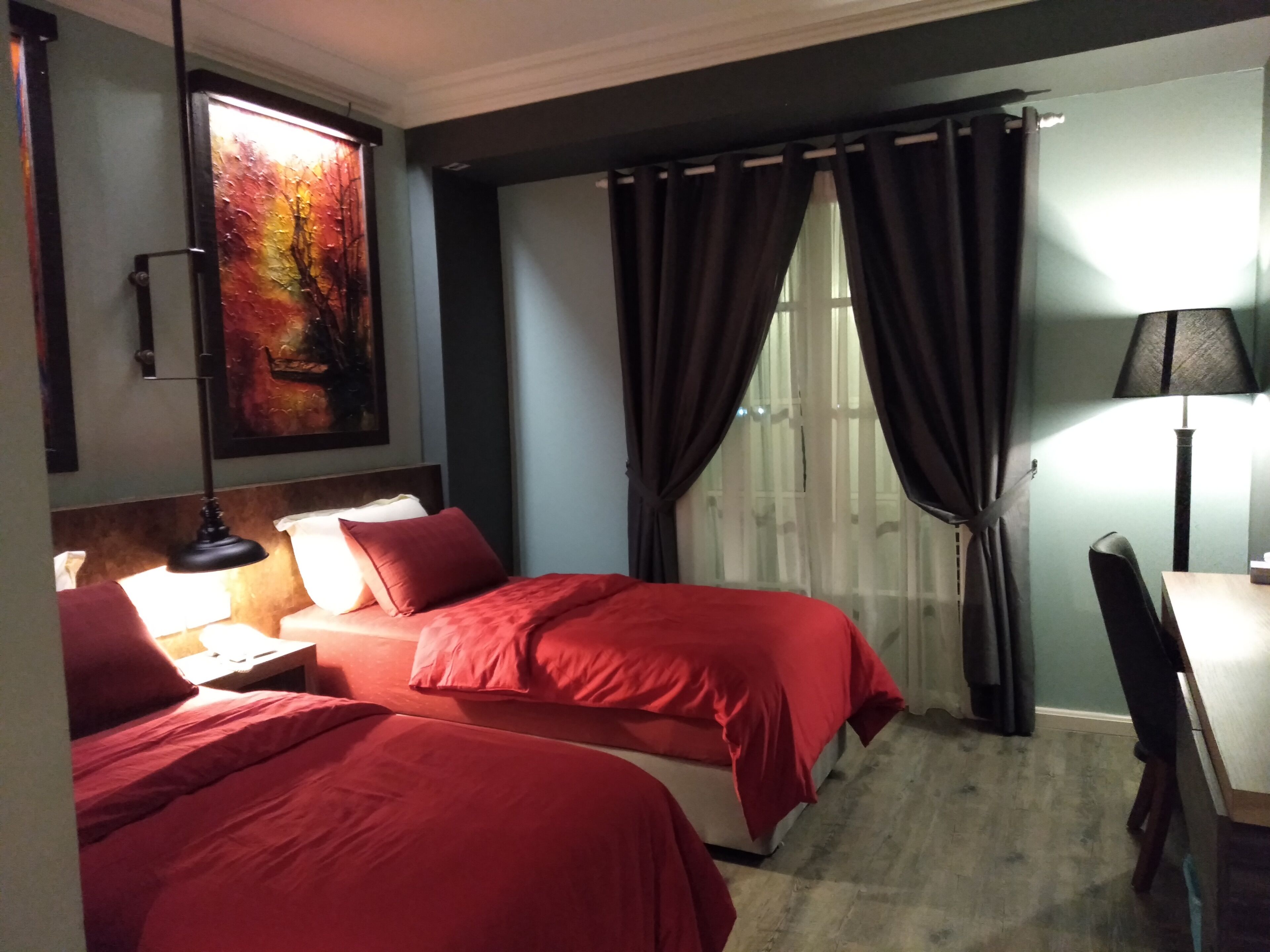 A hotel room with two beds, a desk, a chair, a lamp, and a painting on the wall.
