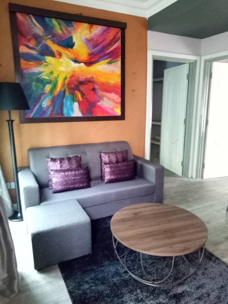 A modern living room with a grey sofa, two purple cushions, a round wooden coffee table, and a colorful abstract painting on the wall.