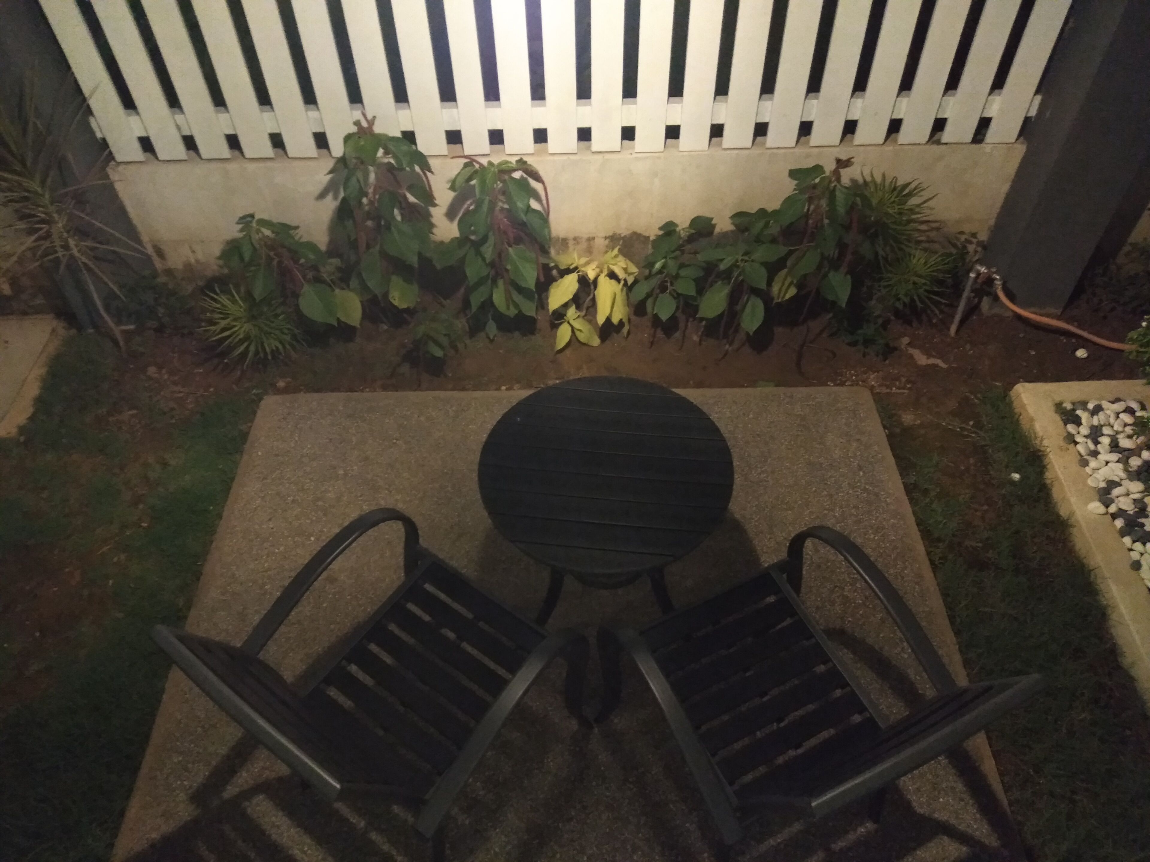 Une terrasse avec une table ronde et deux chaises, entourée de plantes et d’une clôture blanche.