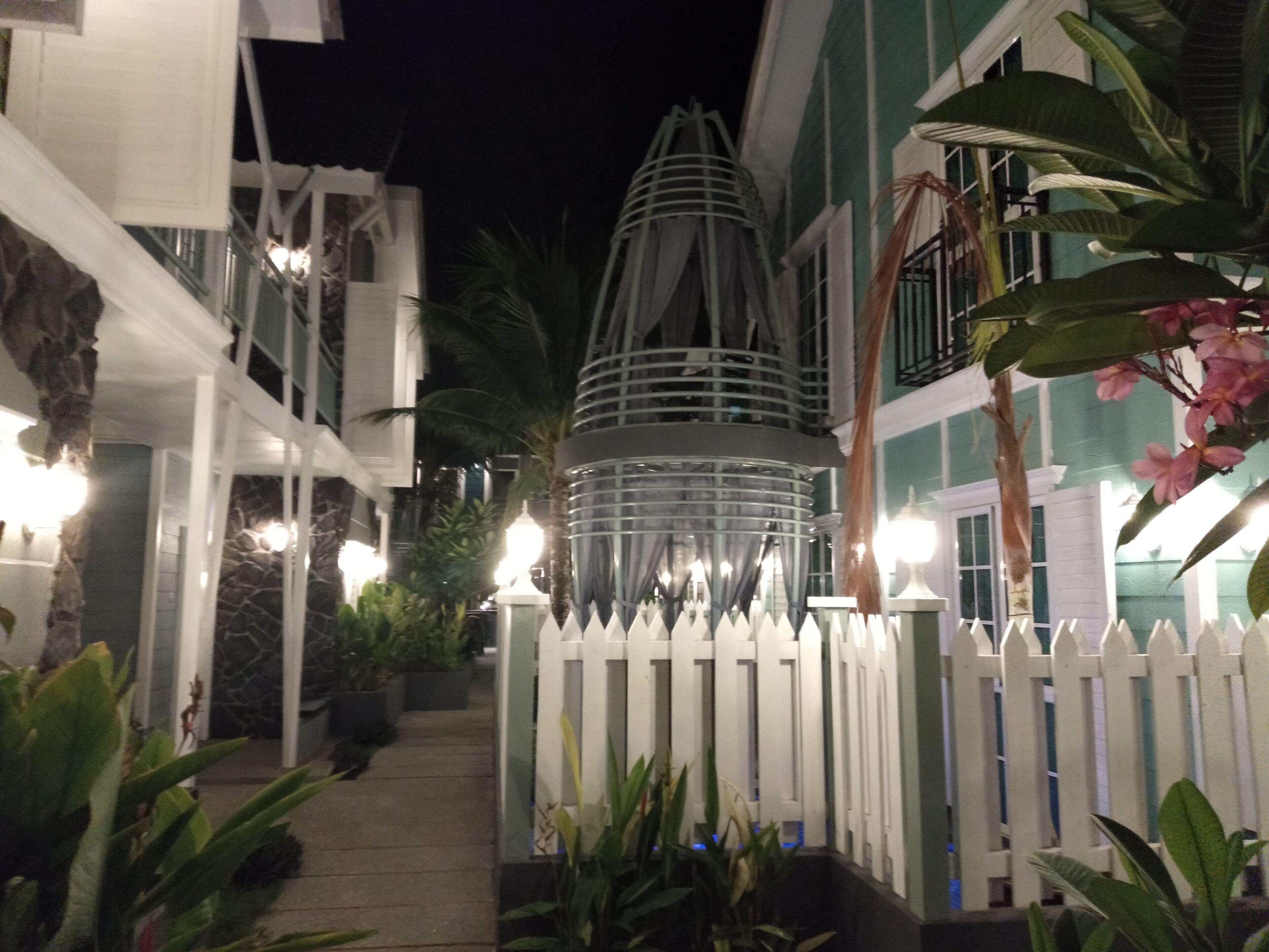 A nighttime view of a hotel with a white picket fence, potted plants, and a decorative metal structure.