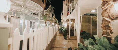 Vista nocturna de un pasillo de hotel con barandillas blancas, pared de piedra y plantas en macetas.