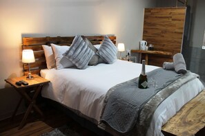 A neatly made bed with a wooden headboard, two bedside lamps, a bottle of champagne, and a small table with a cup and a bottle. - Potties Guest House (Emthanjeni)