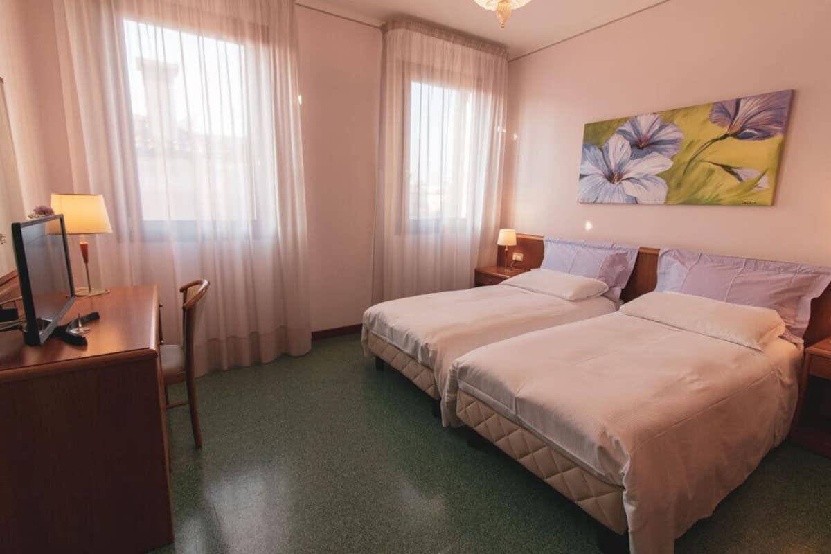 A hotel room with two beds, a desk with a lamp, a painting on the wall, and sheer curtains.
