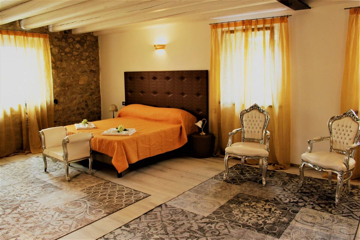 Une chambre d’hôtel avec un grand lit, deux fauteuils décoratifs, une table de chevet et un tapis à motifs.