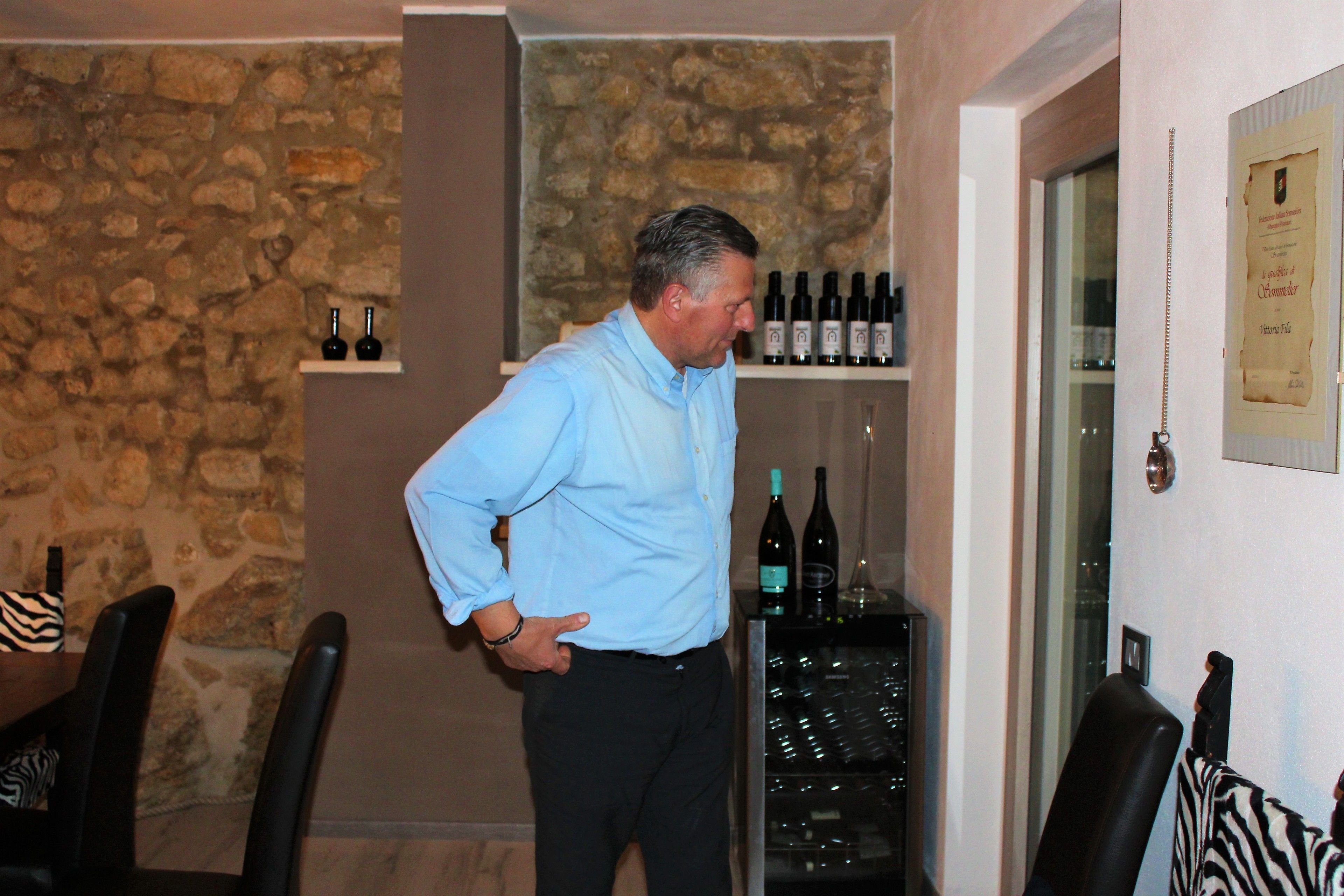A man standing in a room with a stone wall, a wine rack with bottles, and a framed certificate on the wall.