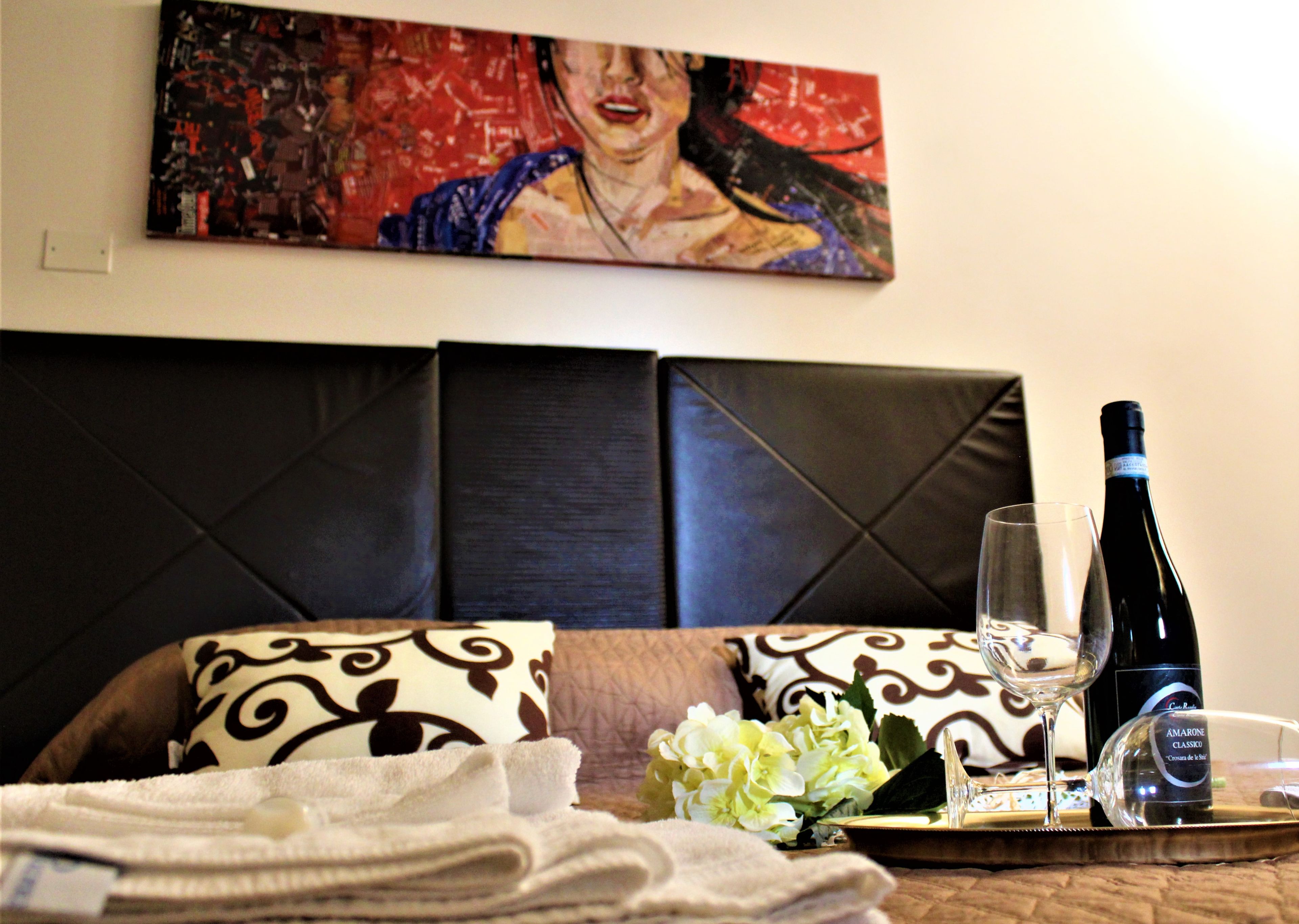 A hotel room with a bed, a painting on the wall, a bottle of wine, a glass, and a tray with flowers and towels.