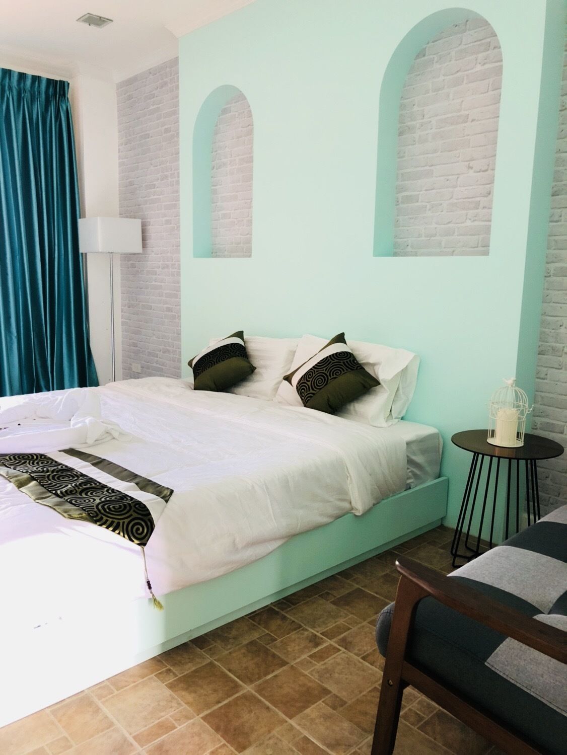 A bed with white bedding and patterned pillows, a small round table with a white candle, and a chair with a striped cushion.