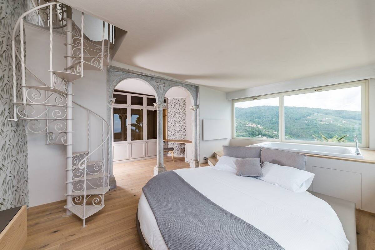 A bedroom with a large bed, a spiral staircase, a view of the outdoors, and a window with a view of the landscape.