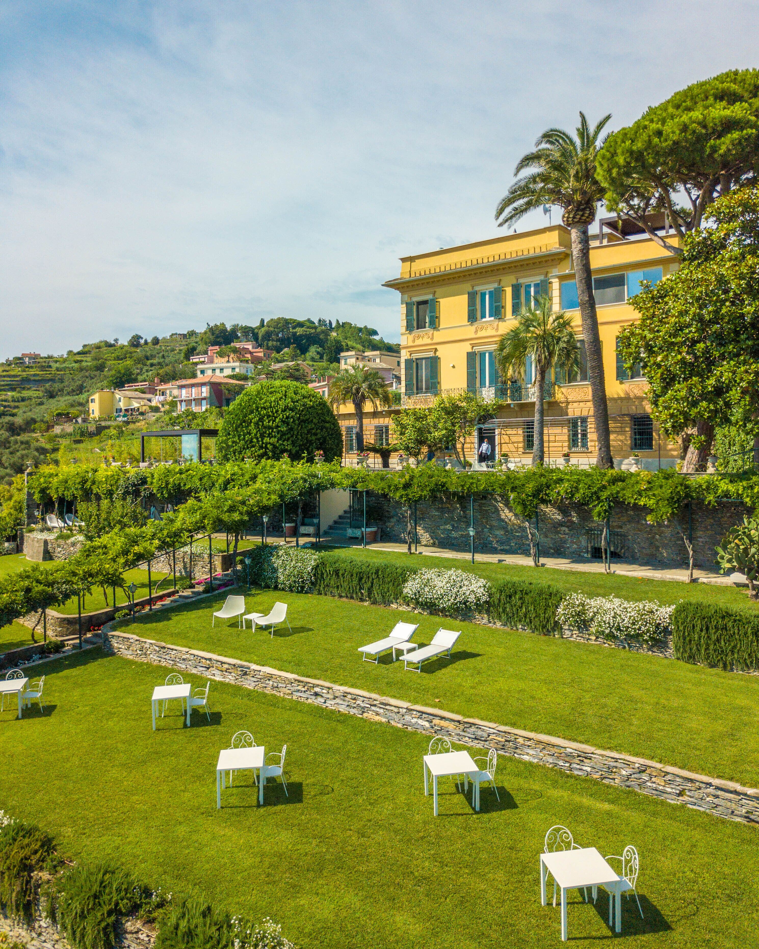A well-maintained garden with white tables and chairs, a stone pathway, and a yellow building with green shutters in the background.