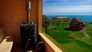 Una cabaña de madera con chimenea de piedra, ventana con vista a un área verde y al mar.
