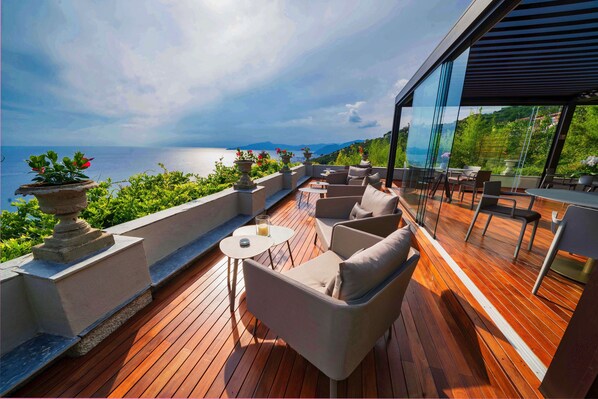 Una terraza de madera con mobiliario exterior, barandilla de cristal y vista al mar.