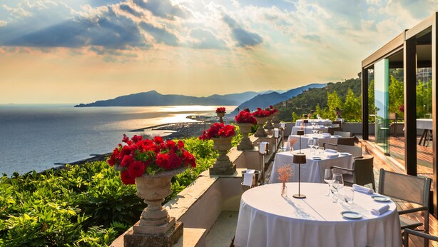 Un restaurante frente al mar con terraza al aire libre, con macetas de piedra con flores rojas y vistas despejadas del mar y montañas lejanas.