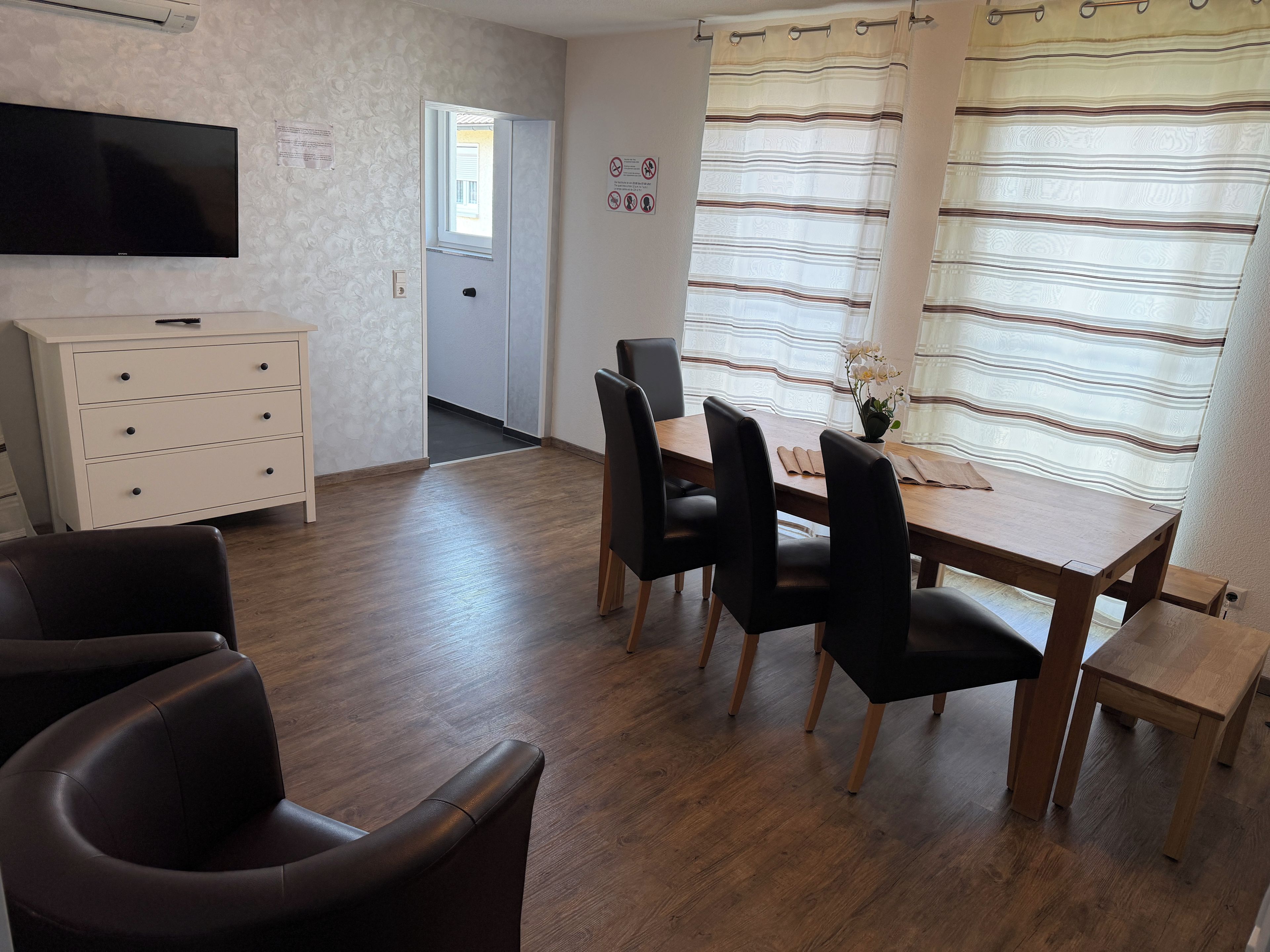 Una habitación con una mesa de comedor de madera, sillas negras, un armario blanco, un televisor de pantalla plana montado en la pared y una ventana con cortinas a rayas.