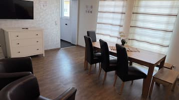 Una habitación con una mesa de comedor de madera, sillas negras, un armario blanco, un televisor de pantalla plana montado en la pared y una ventana con cortinas a rayas.