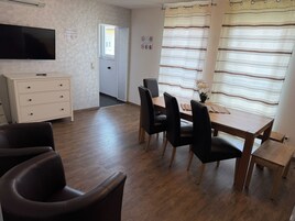 A room with a wooden dining table, black chairs, a white cabinet, a flat-screen TV mounted on the wall, and a window with striped curtains.