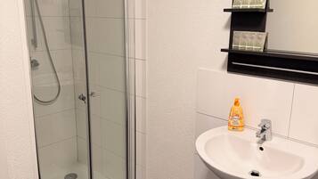 Un baño moderno con ducha envuelta en vidrio, un lavamanos blanco con dispensador de jabón y un espejo con un estante de madera encima.