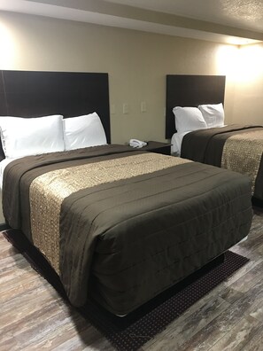 A hotel room with two beds, each with a dark brown bedspread and white pillows. There is a small bedside table with a telephone and a wall-mounted light. The room has a wooden floor and a dark brown rug. - Memphis Inn (Memphis)