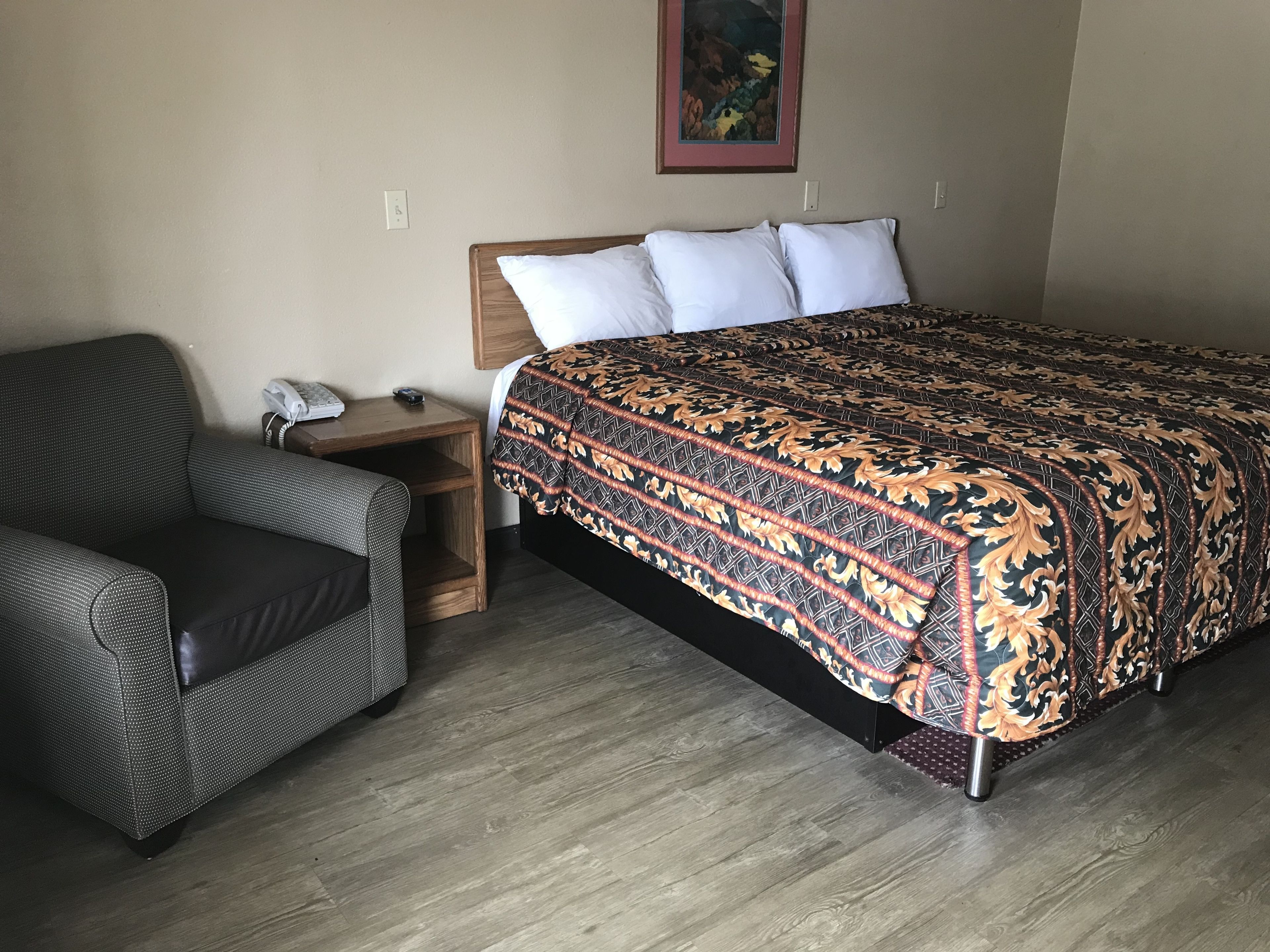 A hotel room with a bed, a chair, a nightstand, and a painting on the wall.