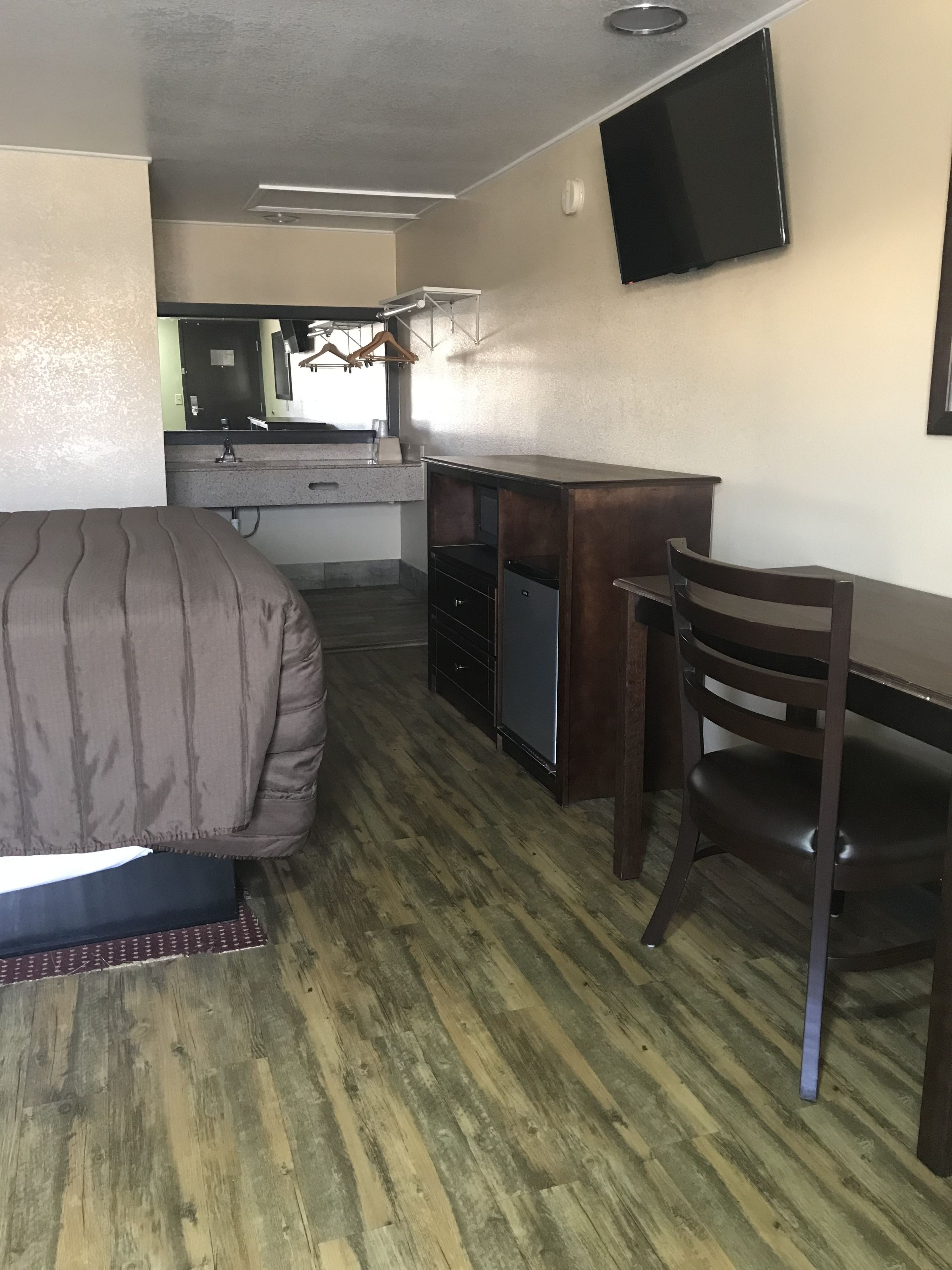 A compact room with a bed, a desk, a chair, a small kitchenette, and a TV mounted on the wall.