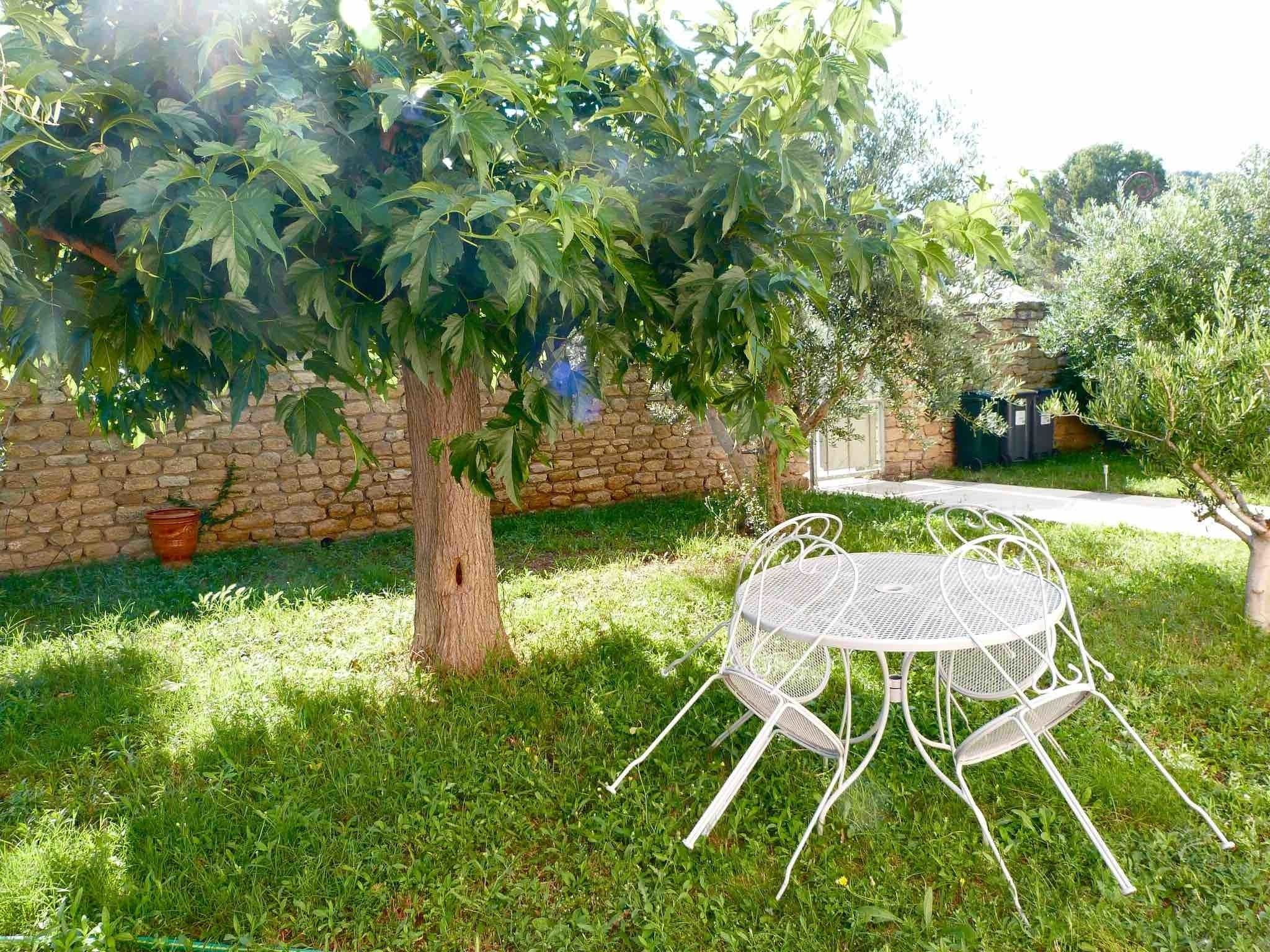 Een terras met een metalen tafel en stoelen, omgeven door gras en een boom.