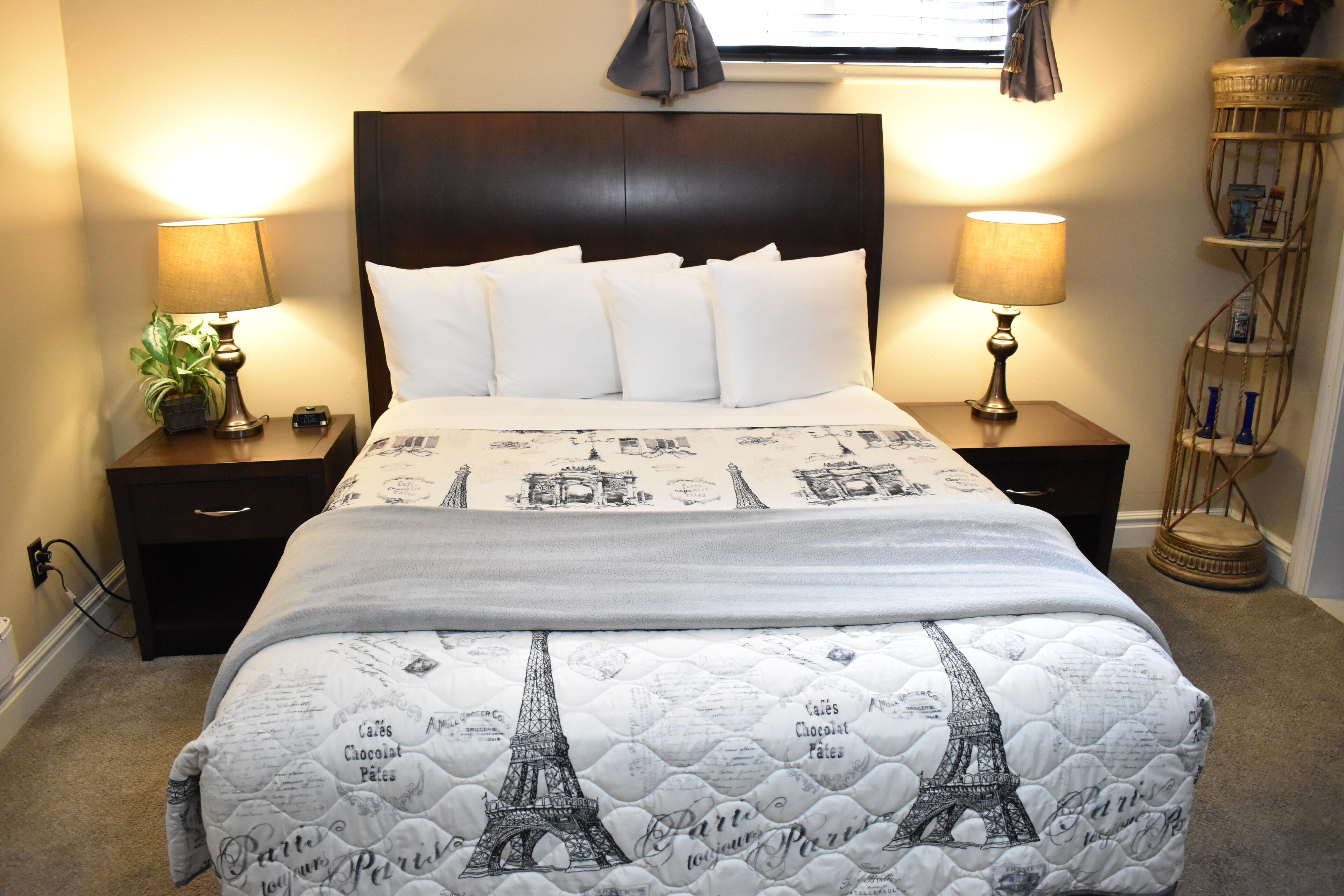 A neatly made bed with a Paris-themed quilt, two bedside lamps, a small plant, and a nightstand with a drawer.