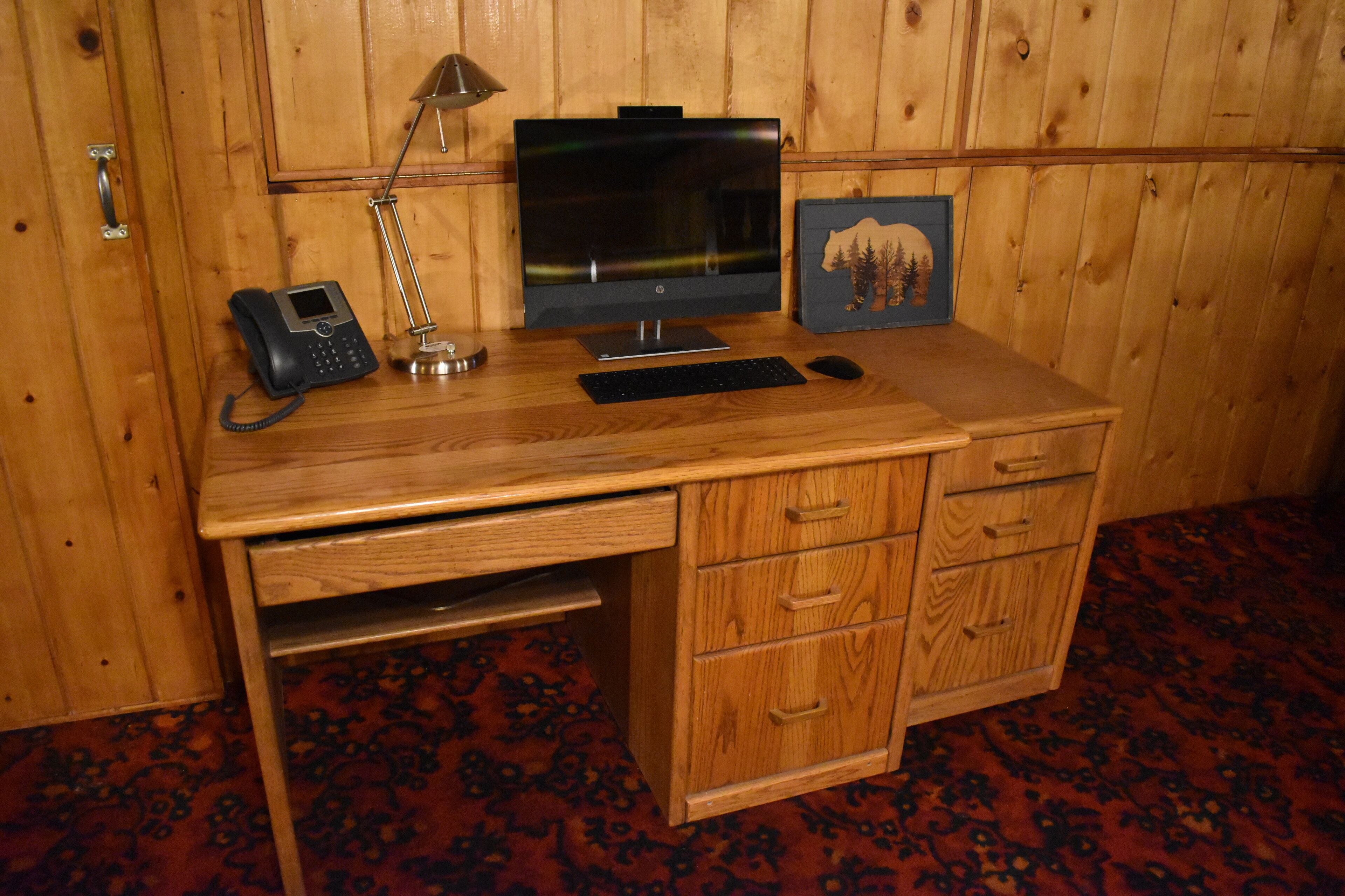 Ein Holztisch mit einem Computer, einem Telefon, einer Lampe und einem gerahmten Bild.