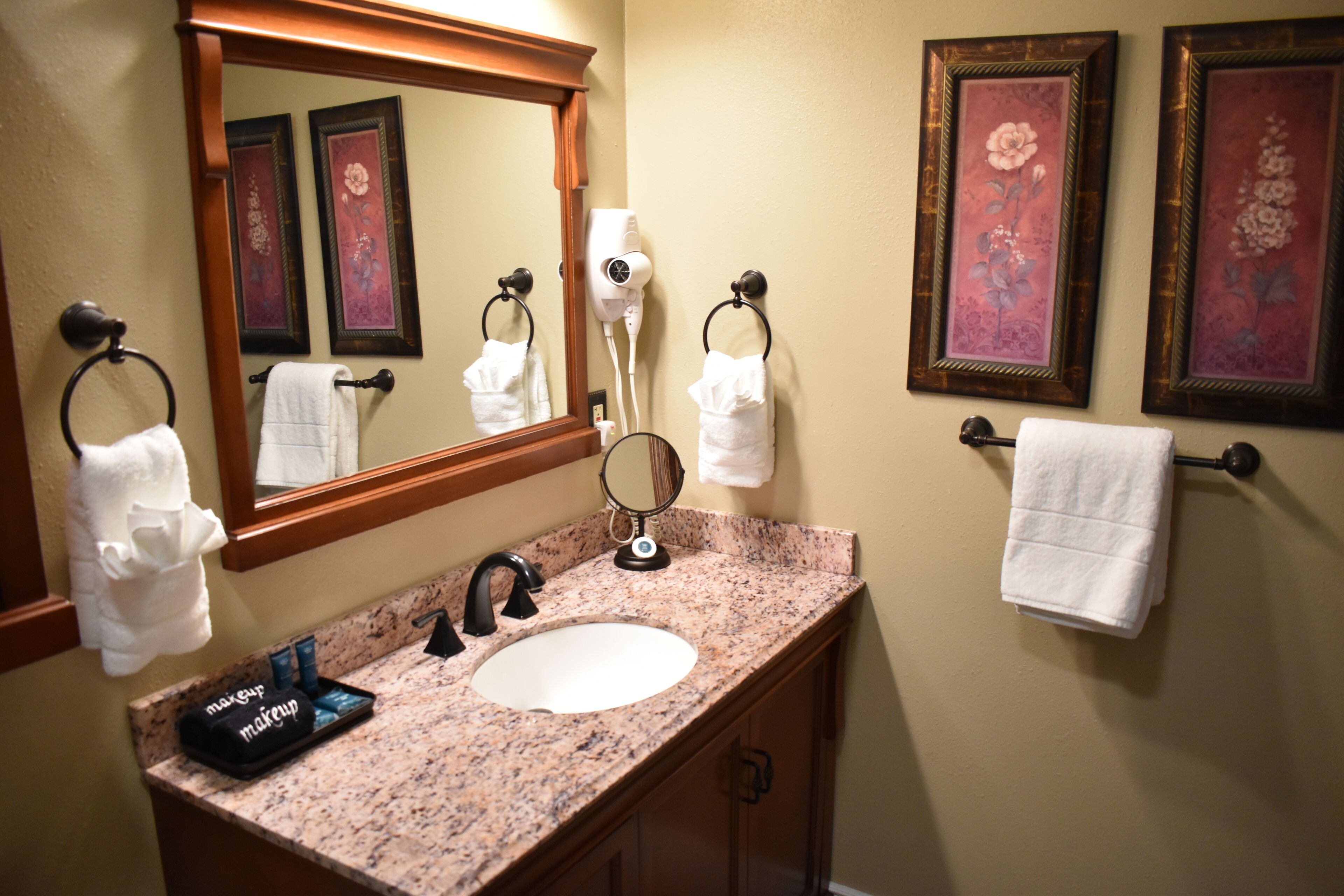 Une salle de bain avec un lavabo doté d’un plan de travail en granit, un miroir et deux tableaux floraux encadrés accrochés au mur.