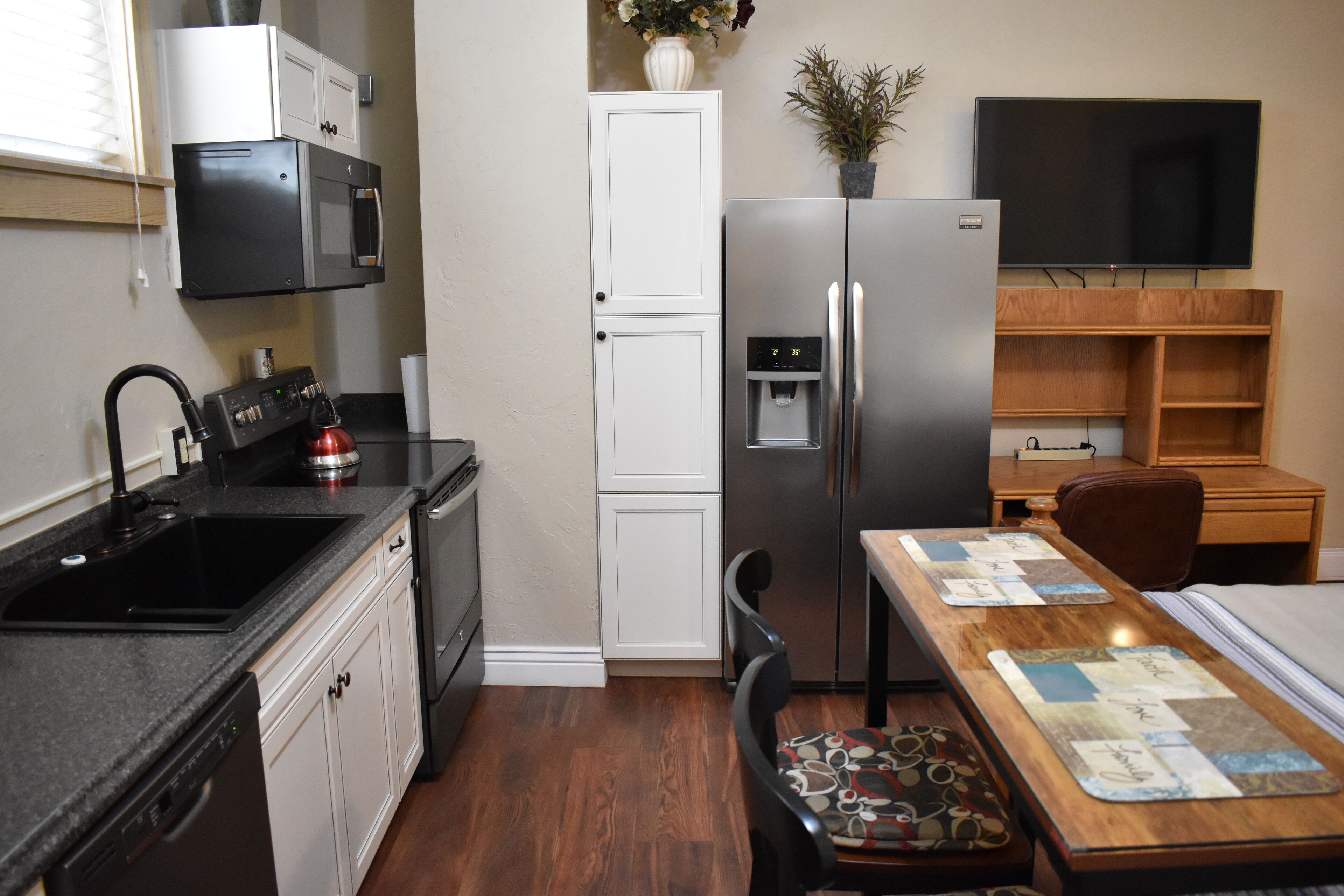 A compact kitchen with a dishwasher, stove, and refrigerator. There is a dining table with chairs and a TV mounted on the wall.