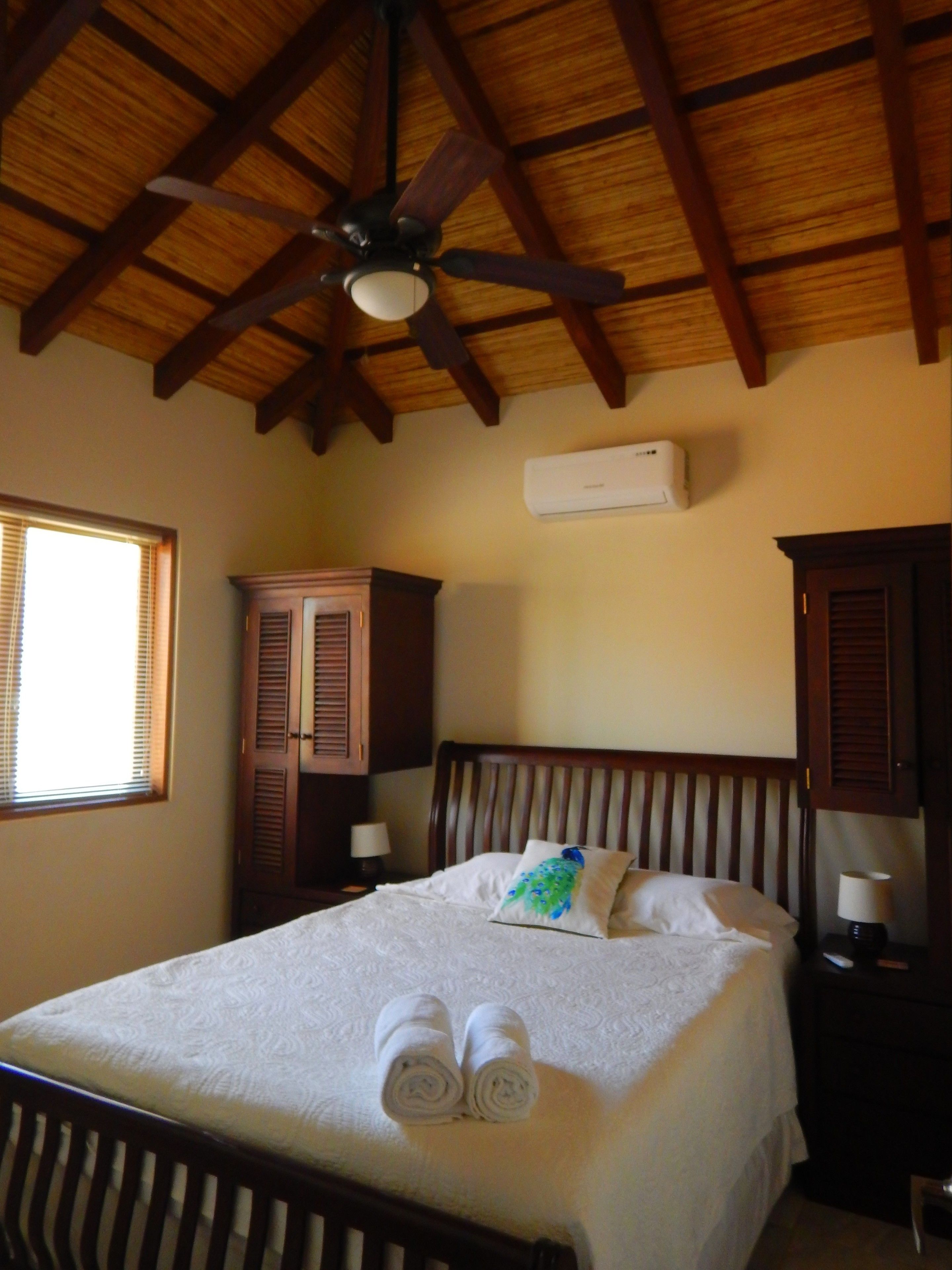 A bedroom with a wooden ceiling, a bed with white linens, two rolled towels, a nightstand with a lamp, and a window with blinds.