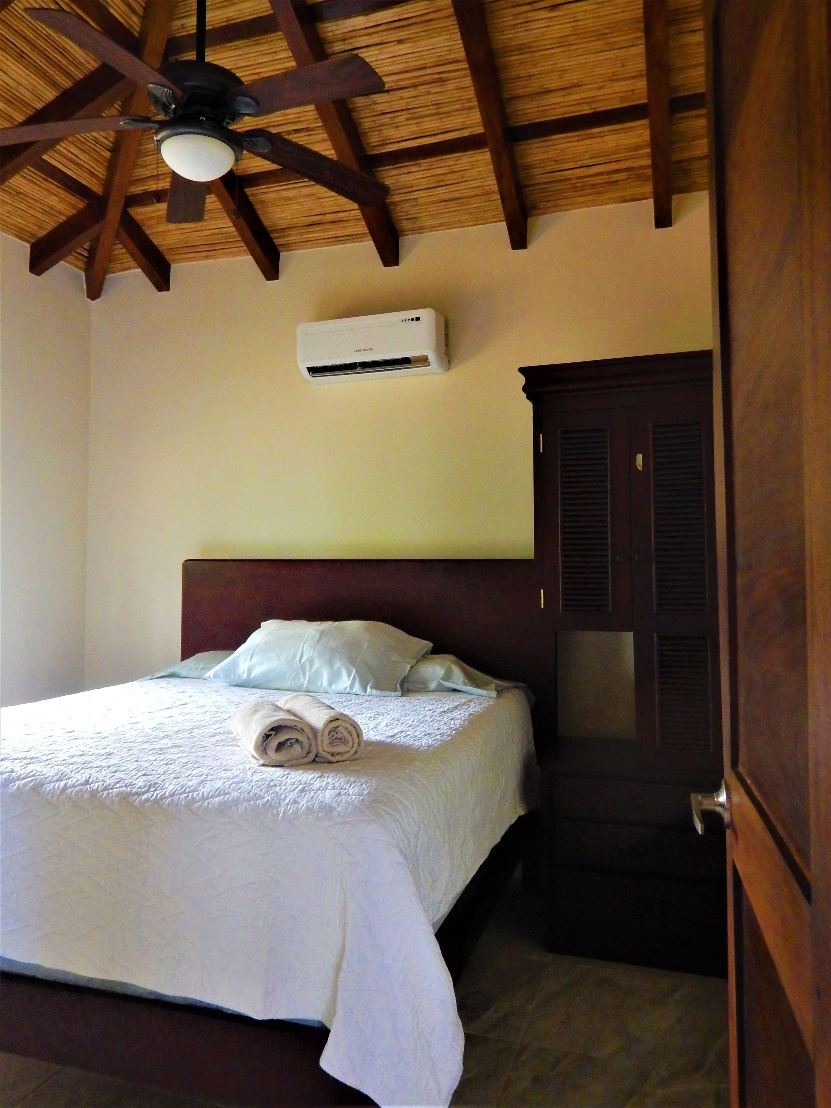 A hotel room with a bed, a ceiling fan, an air conditioning unit, and a wooden wardrobe.